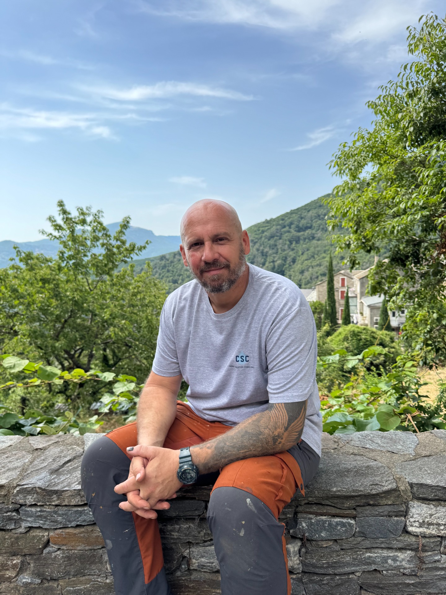 Homme assis sur un muret en pierre, t-shirt gris, montagnes et verdure en arrière-plan.